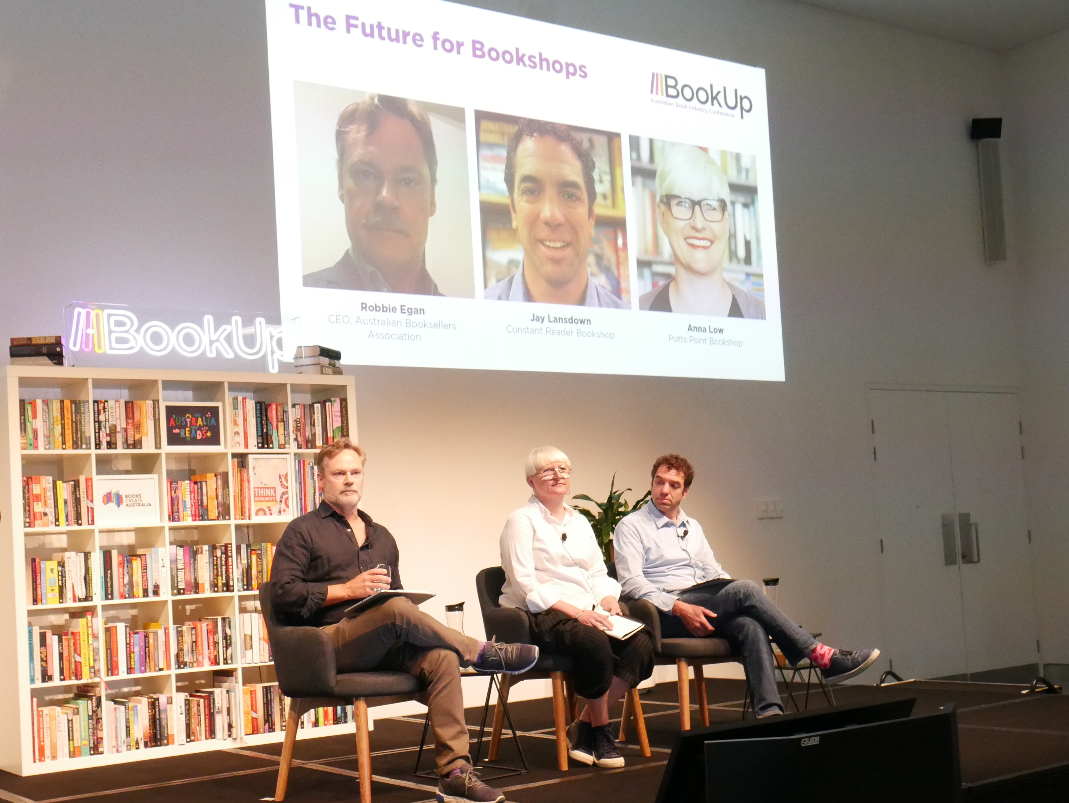 A photograph of two men and a woman, sitting in chairs on the BookUp stage. Projected onto the wall behind them is a slide which reads 'The Future for Bookshops' above headshots of the speakers.