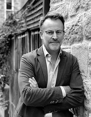 A black and white photograph of James Kellow leaning against a stone wall. James wears glasses and a casual suit, his arms are crossed.