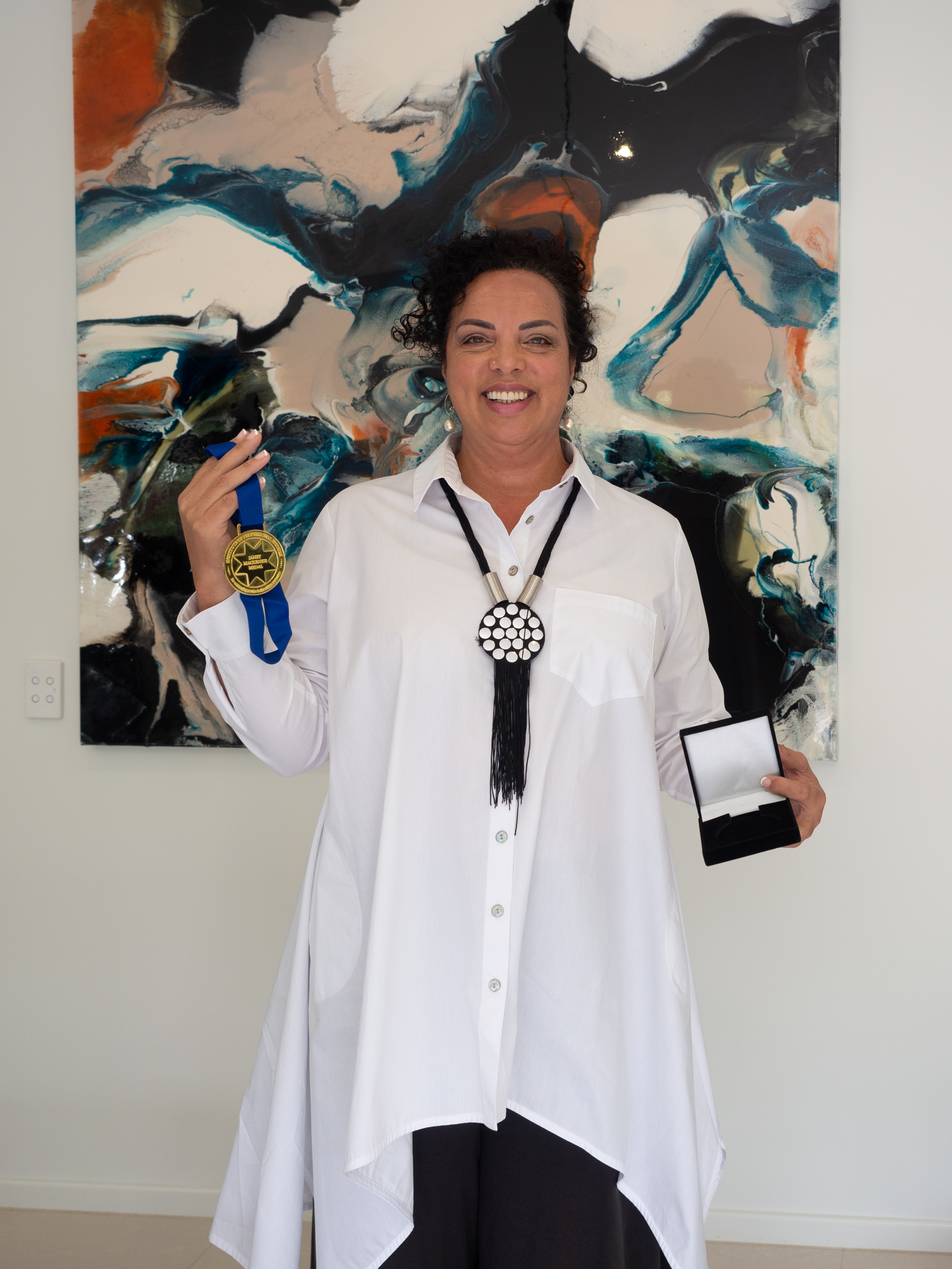 A photo of a smiling woman, Dr Renée Otmar, holding a large gold medal on a blue ribbon, in front of a bright abstract painting