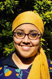 Headshot of Radhiah Chowdhury. Radhiah stands in front of a tree and wears a blue flower print blouse, glasses and a yellow headscarf.