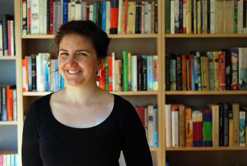 A photograph of Susannah Chambers. Susannah has brown hair, tied back behind her head, and wears a black long sleeve shirt. She stands in front of a row of bookshelves.